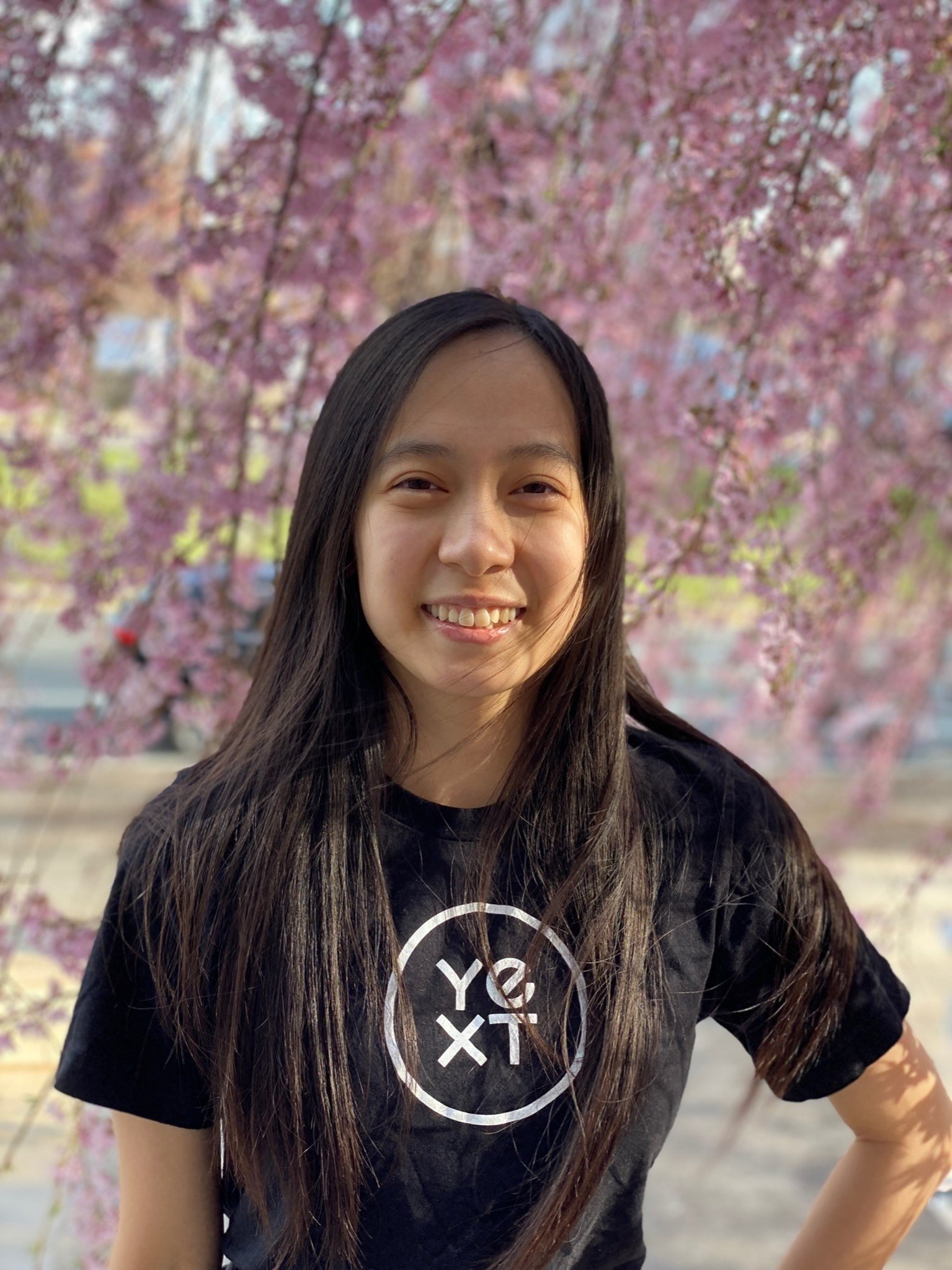 profile picture of Tiffany Trinh, smiling with long black/brown hair, black "YEXT" t-shirt, and pink cherry blossoms blooming in the background